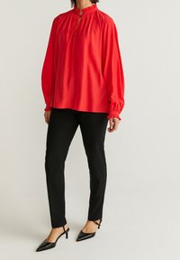 Röd blus med långa ärmar, volangkrage och knyt-detalj, som matchas med svarta slim-fit byxor och spetsiga svarta klackar. Enkel design, slät textur.