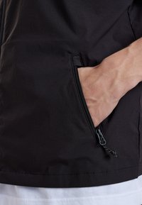 Schwarze Jacke mit einer Reißverschlusstasche, leichtem Stoff, glatter Textur und kontrastierender Naht am Saum, auf einem weißen Hintergrund positioniert.