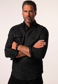 Zwarte button-up shirt met twee borstzakken, lange mouwen opgerold bij de manchetten, en een lichte textuur. Model heeft de armen gekruist voor een neutrale achtergrond.