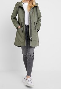 Olivově zelená parka s kapucí, šňůrkami, kapsami na zip a dlouhými rukávy. Nošená přes bílé tričko a šedé džíny, doplněná bílými teniskami.