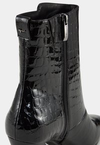 Bottine en cuir verni noir avec texture crocodile, fermeture éclair latérale et petit talon, sur un fond clair uni.