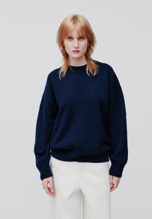 Pull en maille oversize bleu marine avec ourlet et poignets côtelés, avec un col rond et une surface texturée, associé à un pantalon blanc cassé.