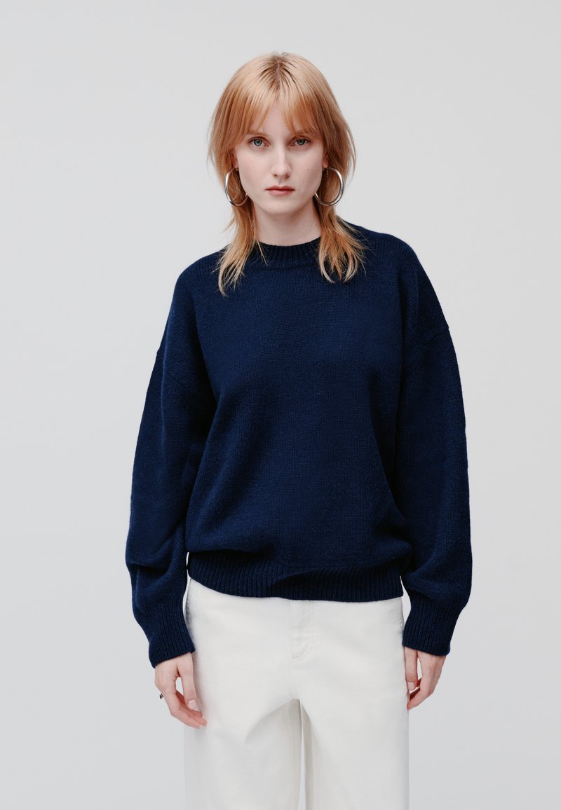 Marineblauwe oversized gebreide trui met geribde zoom en manchetten, voorzien van een ronde hals en een gestructureerd oppervlak, gecombineerd met off-white broeken.
