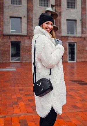 Weißer, flauschiger, übergroßer Mantel, schwarze Umhängetasche mit geometrischem Design und graue Handschuhe vor einem Backstein-Hintergrund.