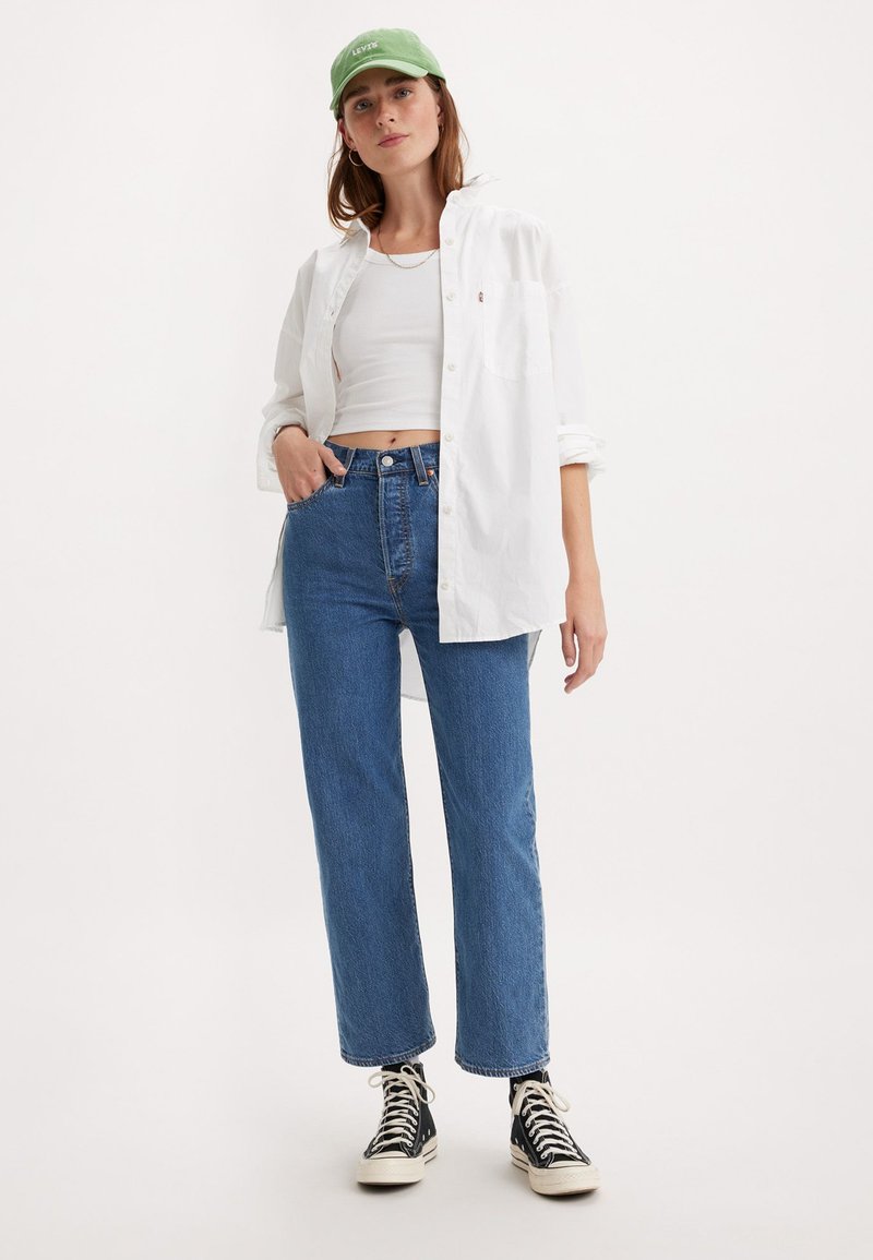 Chemise blanche à manches longues sur un crop top blanc, associée à un jean bleu taille haute et des baskets montantes noires. Casquette verte avec logo.