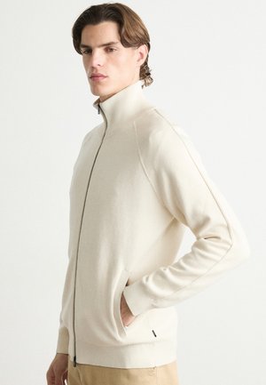 Maglione beige con zip, colletto alto, polsini a coste e tasche laterali, realizzato in un tessuto morbido, con dettagli cuciti lungo le spalle.