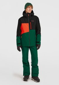 Groene en zwarte skijas met oranje accenten, met ritszakken en een capuchon. Gecombineerd met bijpassende groene broek en zwarte laarzen.