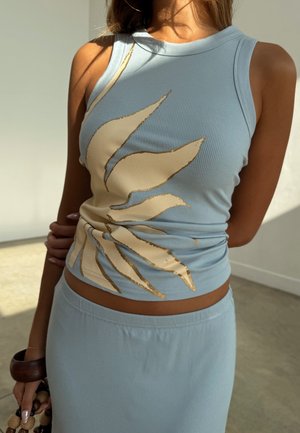 Femeie purtând un set format din top fără mâneci și fustă albastru deschis, cu dungi și design de frunze bej, ținând o geantă cu mărgele și o brățară din lemn într-o cameră luminată de soare.