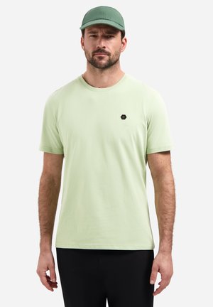 Man draagt een lichtgroen T-shirt met een klein zwart logo op de borst, een bijpassende groene pet en zwarte broek, staand tegen een witte achtergrond.