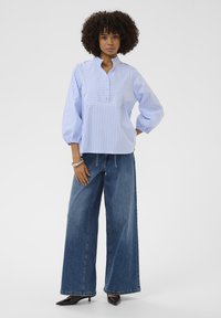 Bluză cu mâneci lungi cu dungi albastre deschise și albe, cu guler drept și fâșie frontală cu nasturi, asortată cu blugi evazați din denim închis la culoare și pantofi cu toc negri.