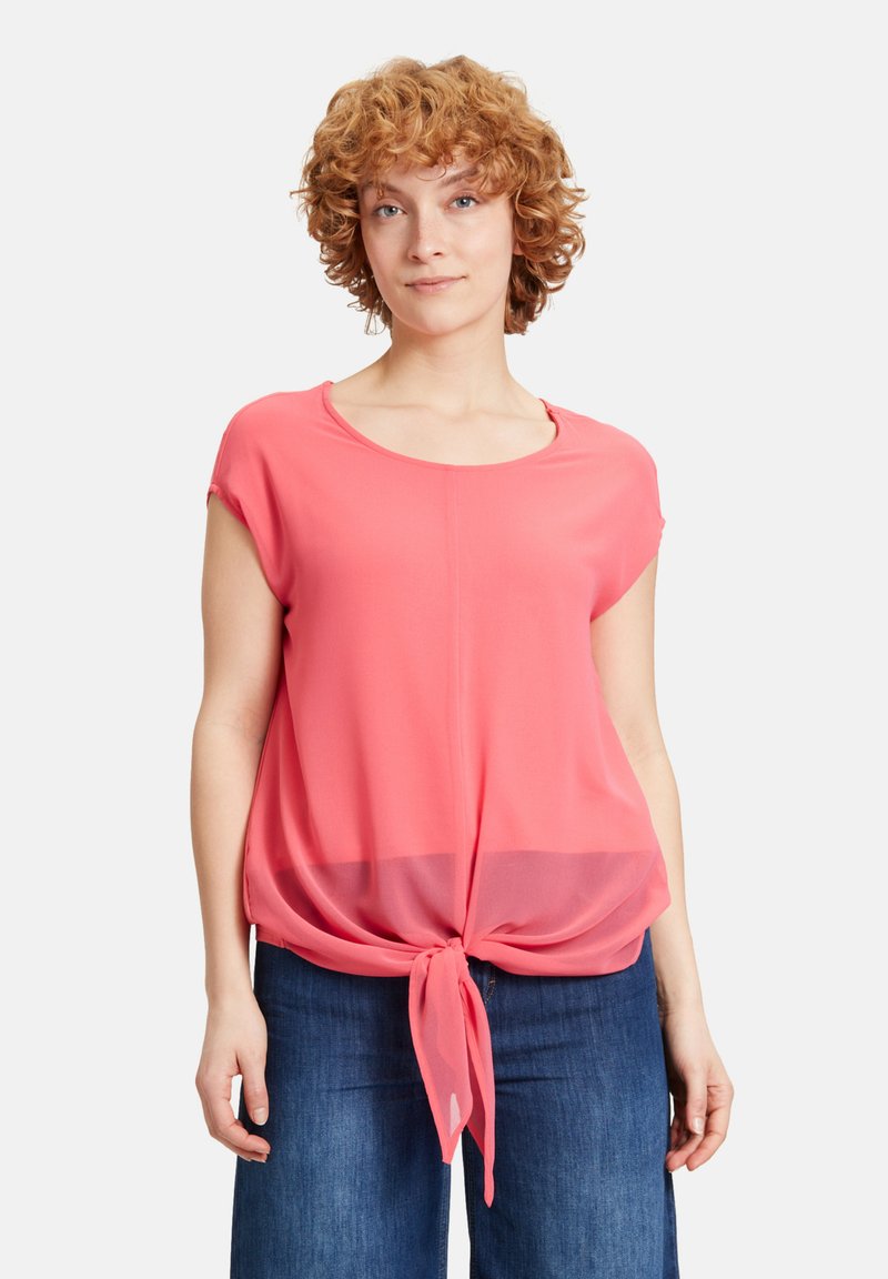 Top corto a maniche corte di colore corallo, realizzato in tessuto leggero, con un dettaglio a nodo frontale e un collo rotondo. Abbinato a pantaloni in denim.