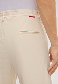 Beige broek met elastische taille en een achterzak, gemaakt van gladde stof. Voorzien van een klein rood label met de tekst "SOFT JEANS".