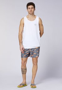Weißes ärmelloses Tanktop mit kleinem Logo; graue Badeshorts mit orangefarbenem Känguru-Muster; graue Flip-Flops mit gelber Sohle.