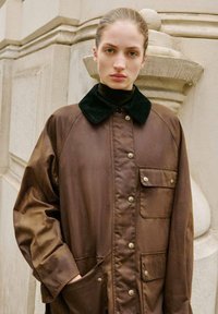 Brun, letvægts yderjakke med en sort fløjlskrave, trykknapper og forsække. Glat tekstur og afslappet pasform.