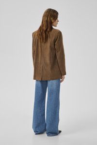 Sieviete ar gariem brūniem matiem, valkājot brūnu zamšādas žaketi un plašu zilas džinsa bikses, stāvot ar muguru pret vienkāršu fonu.