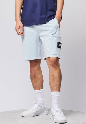 Mann trägt hellblaue Cargoshorts, weiße Sportsocken, weiße Turnschuhe und ein dunkelblaues T-Shirt, steht mit den Händen in den Taschen.