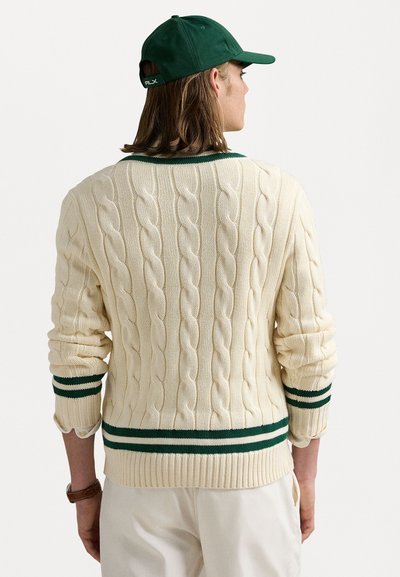 Creme farvet kabelstrikket sweater med grønne striber i krave og manchetter, der har lange ærmer og ribbet kant. Tilbehøret inkluderer en grøn cap.