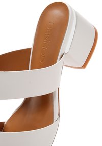 Sandales à talons blocs blanches en matériau synthétique lisse, avec des brides larges, une semelle intérieure brune et une légère courbure au niveau du talon. Nom de la marque visible.