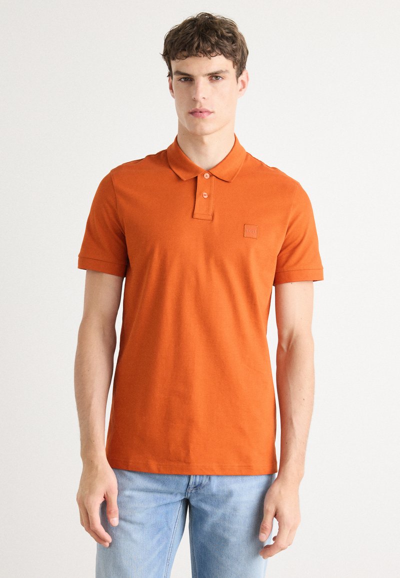 Oransje bomullspolo med klassisk krage, korte ermer, brodert logo på brystet og rett nederkant. Brukt sammen med blå jeans.