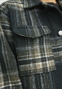 Rutigt yllejacka i mörkgrön och beige, med stora framfickor, texturerad tyg och metallknappsomslag.