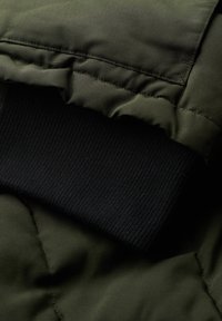 Telaje de chaqueta acolchada verde oscuro con costuras acolchadas; puño negro ajustado de canalé visible, enfatizando la textura y los colores en contraste.