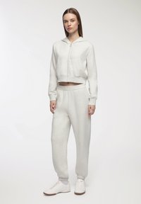 Felpa corta grigia chiara con zip e tasche frontali abbinata a pantaloni della tuta coordinati. Entrambi hanno una texture morbida e una cintura elasticizzata. Sneakers bianche.