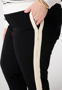 Zwarte broek met een zijpaneel van mesh in beige. De broek heeft een gladde textuur, een aansluitend ontwerp en een zichtbaar zakje aan de zijkant.