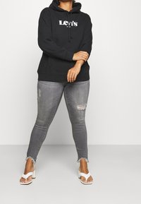 Persona che indossa un hoodie nero di Levi's, jeans skinny strappati grigi e sandali con tacco alto bianchi intrecciati, in piedi con le braccia incrociate.