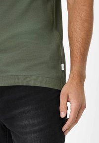 Grøn bluse med tekstur og ribbet kant, delvist stukket ned i sorte jeans. Lille mærke synligt på skjortens side. Hænderne hviler på hoften.