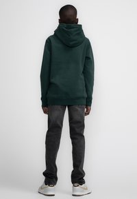 Vihreä huppari hupulla, yhdistettynä tummanharmaisiin farkkuihin. Pehmeä rakenne, rento istuvuus ja yksinkertainen muotoilu ilman erottuvia kuvioita tai yksityiskohtia.