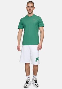 T-shirt di cotone verde con maniche corte e colletto rotondo, abbinato a pantaloni corti bianchi con logo Fila verde. Calzature comprendono sneaker bianche e nere.