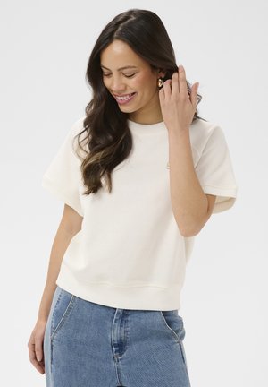 Jeune femme aux longs cheveux noirs portant un haut blanc à manches courtes et un jean bleu, souriante en se touchant les cheveux.