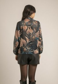Blouse noire transparente avec imprimé floral dans des tons de beige et de bleu, manches longues et coupe décontractée, associée à un short en cuir noir.