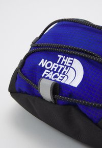 Sac North Face bleu et noir avec logo blanc brodé et cordon noir sécurisé par une boucle grise sur un fond clair.