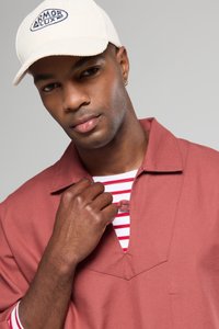 Rode shirt met een open plooi over een wit en rood gestreept onderhemd, in combinatie met een beige pet met een logo.