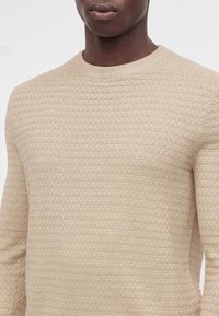 Gros plan d'une personne portant un pull en maille beige texturé avec un col rond, montrant le haut du torse et la zone de la bouche.