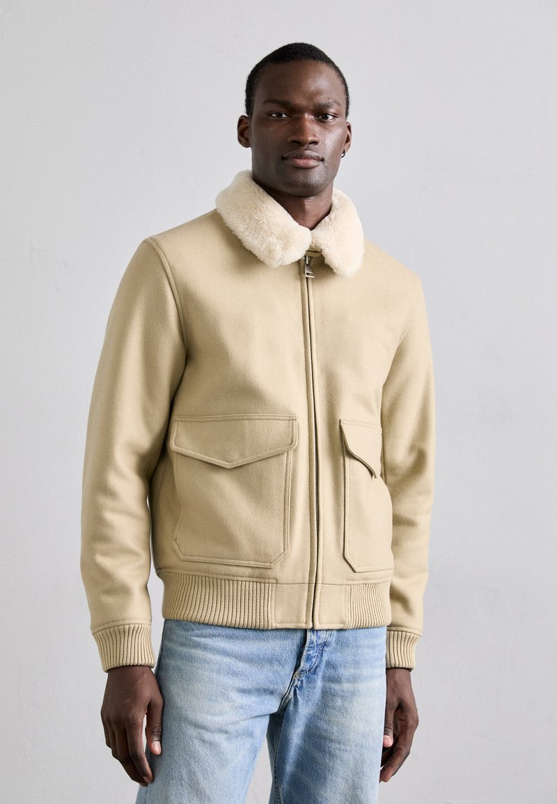 Beige wollen jack met een zachte nepbont kraag, twee voorzakken, geribbelde manchetten en zoom, ritssluiting en een losse pasvorm.