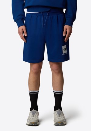 Person trägt blaue Kordelzug-Shorts mit The North Face-Logo, schwarze Socken mit weißen Streifen und hellgraue Wanderschuhe und steht mit entspannten Händen.