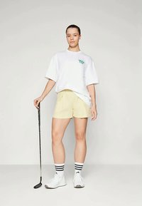 Γυναίκα που στέκεται κρατώντας ένα γκολφ μπάσο, ντύμενη με ένα άσπρο oversized t-shirt, μπεζ σορτς, ριγέ κάλτσες και άσπρα αθλητικά παπούτσια, σε ανοιχτό φόντο.