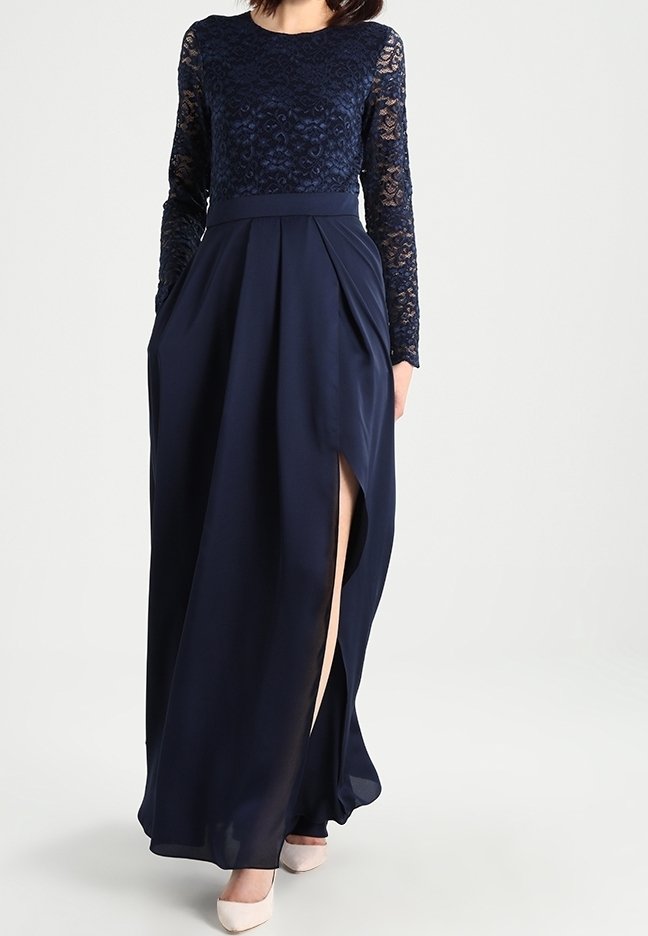 Femme portant une robe top à manches longues en dentelle bleu marine avec une fente haute et des poches, associée à des talons blancs à bout pointu.
