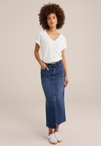 Top bianco a V, con texture, abbinato a una gonna maxi in denim blu con uno spacco frontale. Indossato con sneakers bianche e una cintura nera.