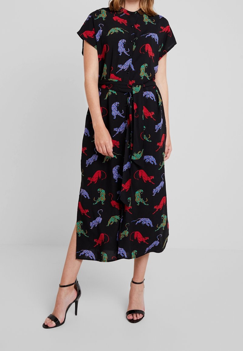 Femme portant une robe midi noire à motifs de léopards colorés rouge, vert et bleu, associée à des sandales noires à talons hauts et bride à la cheville.
