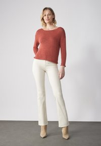 Rustfarbenes gestricktes Sweatshirt mit weitem Ausschnitt, langen Ärmeln und strukturiertem Finish, kombiniert mit weißen Schlagjeans und beigen Knöchelstiefeln.