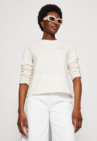 Camicia a maniche lunghe beige chiaro con un fit comodo e maniche arrotolate. Abbinata a pantaloni bianchi. Accessoriata con occhiali da sole oversize con motivo.