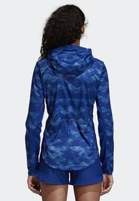 Veste de sport bleue à capuche, avec un motif en zigzag et un tissu léger. Détail comprenant un cordon de serrage à la taille et des manches longues.