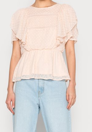 Femme portant un chemisier léger rose clair transparent à volants avec des pois, associé à un jean en denim bleu clair taille haute.