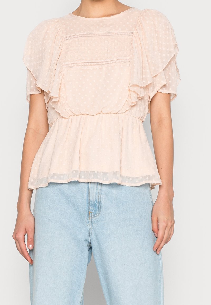 Femme portant un chemisier léger rose clair transparent à volants avec des pois, associé à un jean en denim bleu clair taille haute.