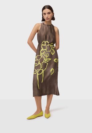 Kobieta w brązowej, bezrękawnej sukience midi z żółtym motywem kwiatowym, w żółtych, szpiczastych balerinach, stoi na jednolitym tle.