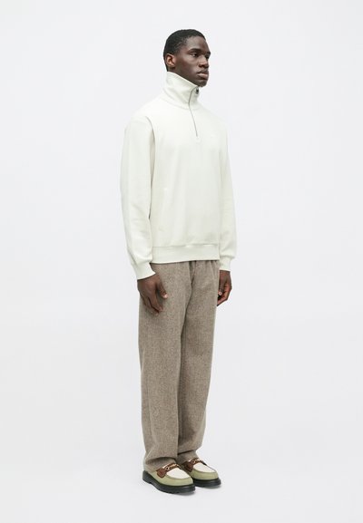 Άνδρας φορεί λευκό πουλόβερ με μισό φερμουάρ, καφέ υφασμάτινα φαρδιά παντελόνια και μπεζ και καφέ loafers, στέκεται μπροστά σε έναν άσπρο φόντο.