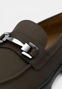 Hnedá kožená mokasína s lesklým strieborným kovovým doplnkom cez priehlavok a jemným prešívaním pozdĺž okrajov. Hladká matná textúra.